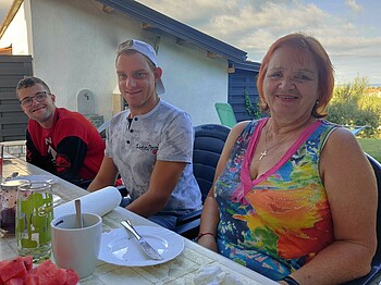 Frau mit roten halblangen Haaren und links davon zwei junge Männer sitzen am Tisch