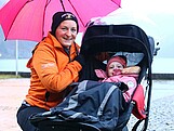 Eine Frau kniet mit Regenschrim bei einem Kind im Kinderwagen.