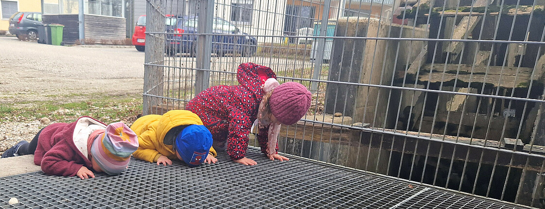 Drei Kinder liegen am Bauch und schauen durch ein Gitter am Boden.
