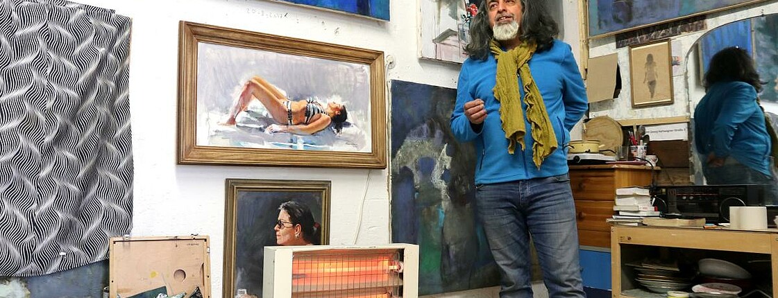 Ein Mann steht in seinem Atelier vor seinen Bildern, die an der Wand hängen.
