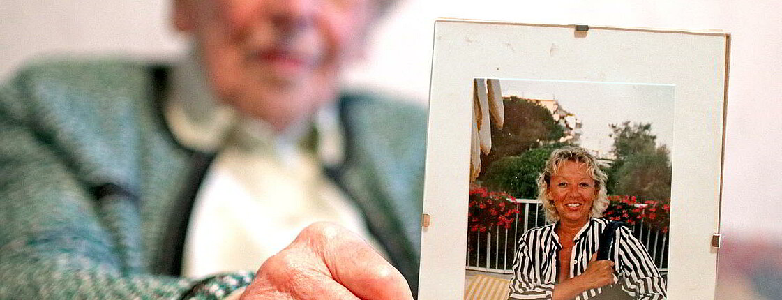 Eine ältere Frau sitzt bei einem Tisch und hält eine Foto ihrer Tochter in den Händen.