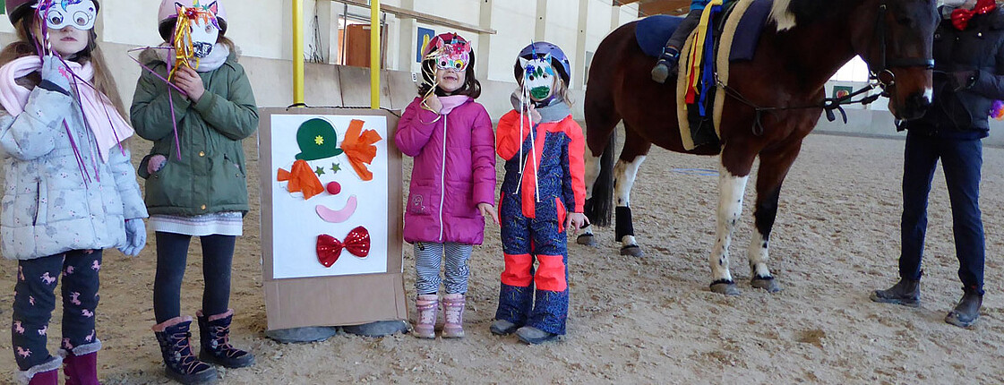 Kinder stehen in der Reithalle mit Einhornmasken und ein geschücktes Pferd steht daneben.